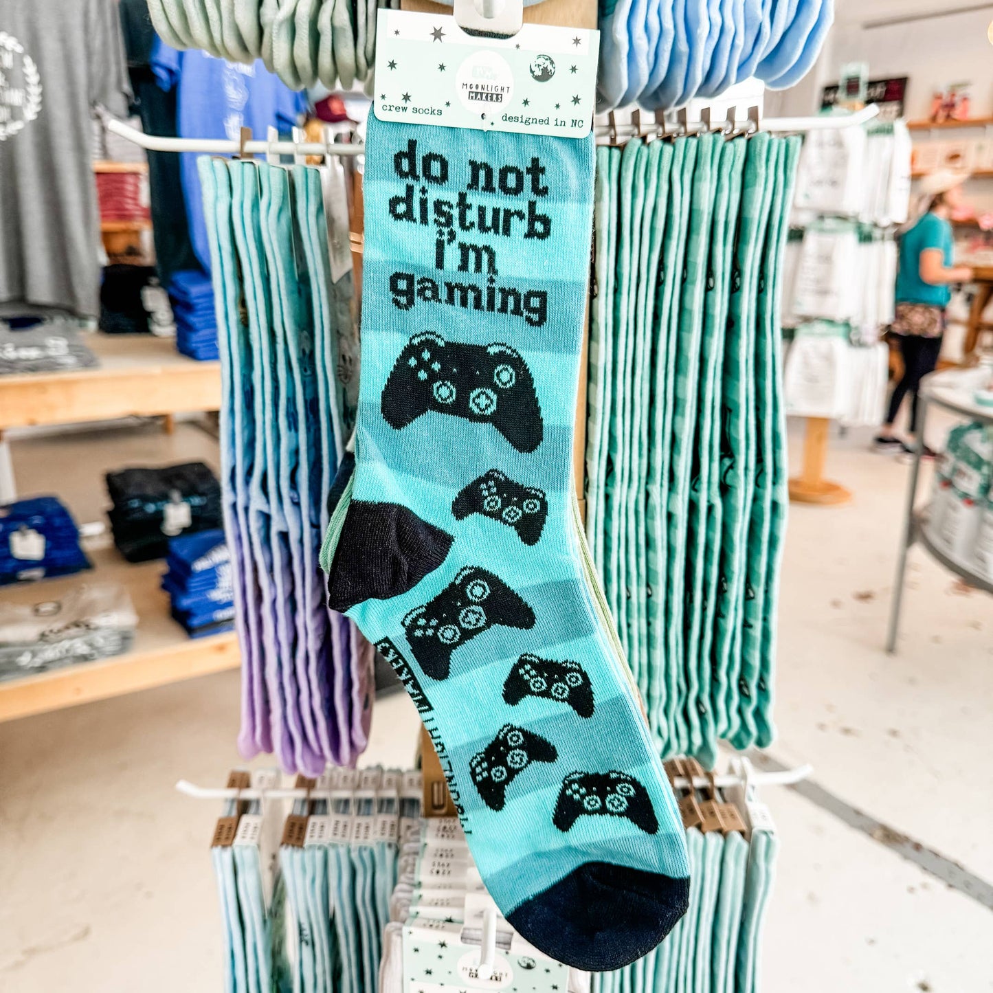 Do Not Disturb I’m Gaming blue crew socks with black video game controller design hanging on a retail display rack inside a gift shop.