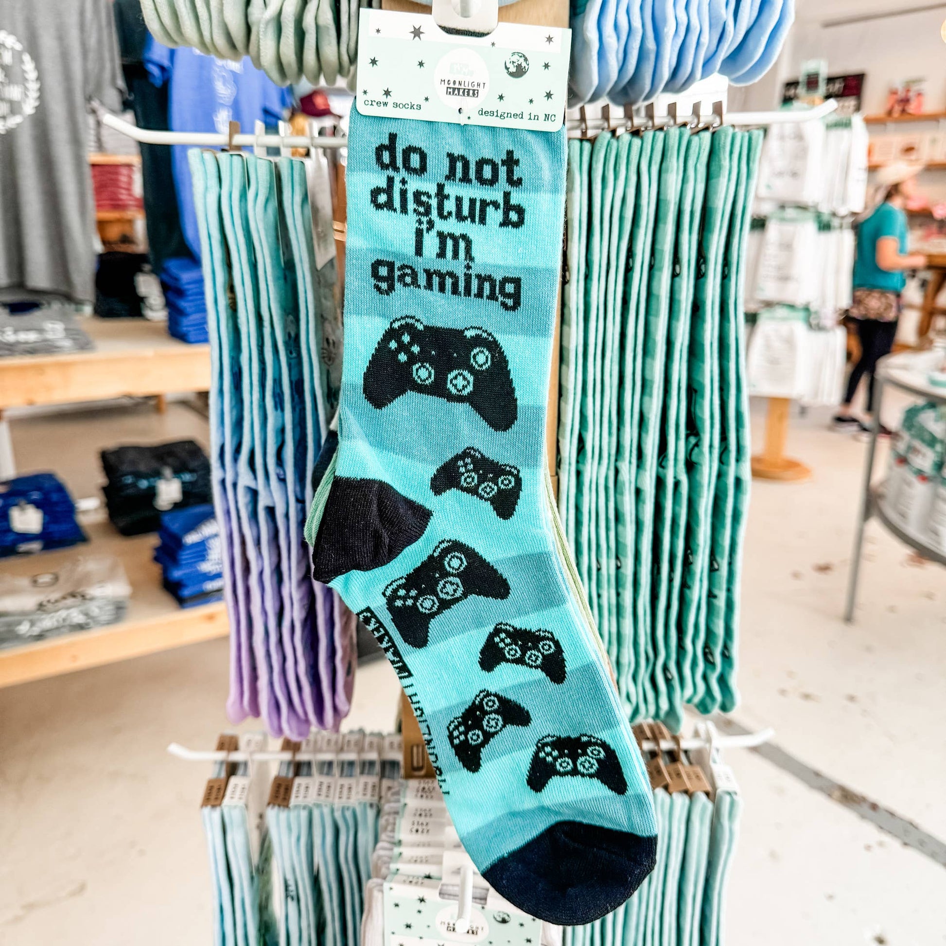 Do Not Disturb I’m Gaming blue crew socks with black video game controller design hanging on a retail display rack inside a gift shop.