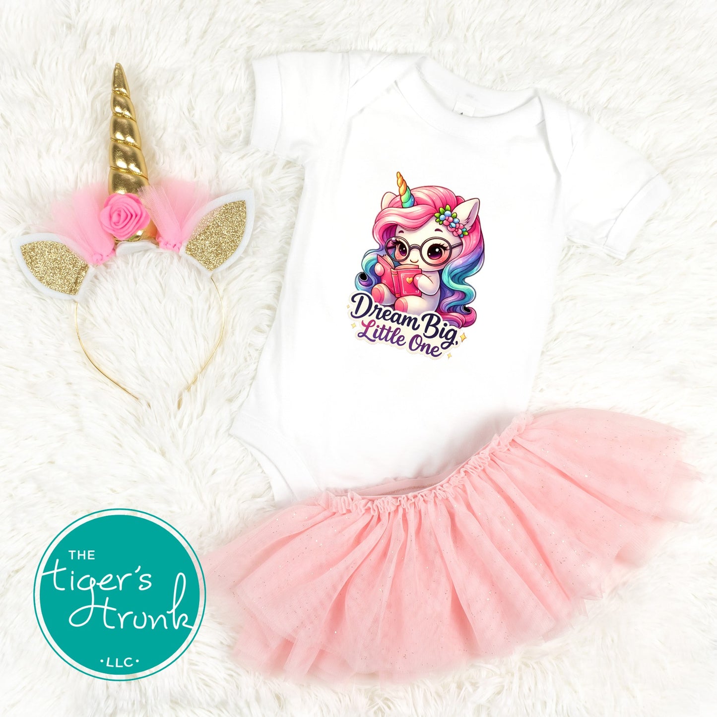 Infant white bodysuit on a white fur background with a colorful unicorn graphic that reads Dream Big Little One next to a tutu and a gold unicorn headband.