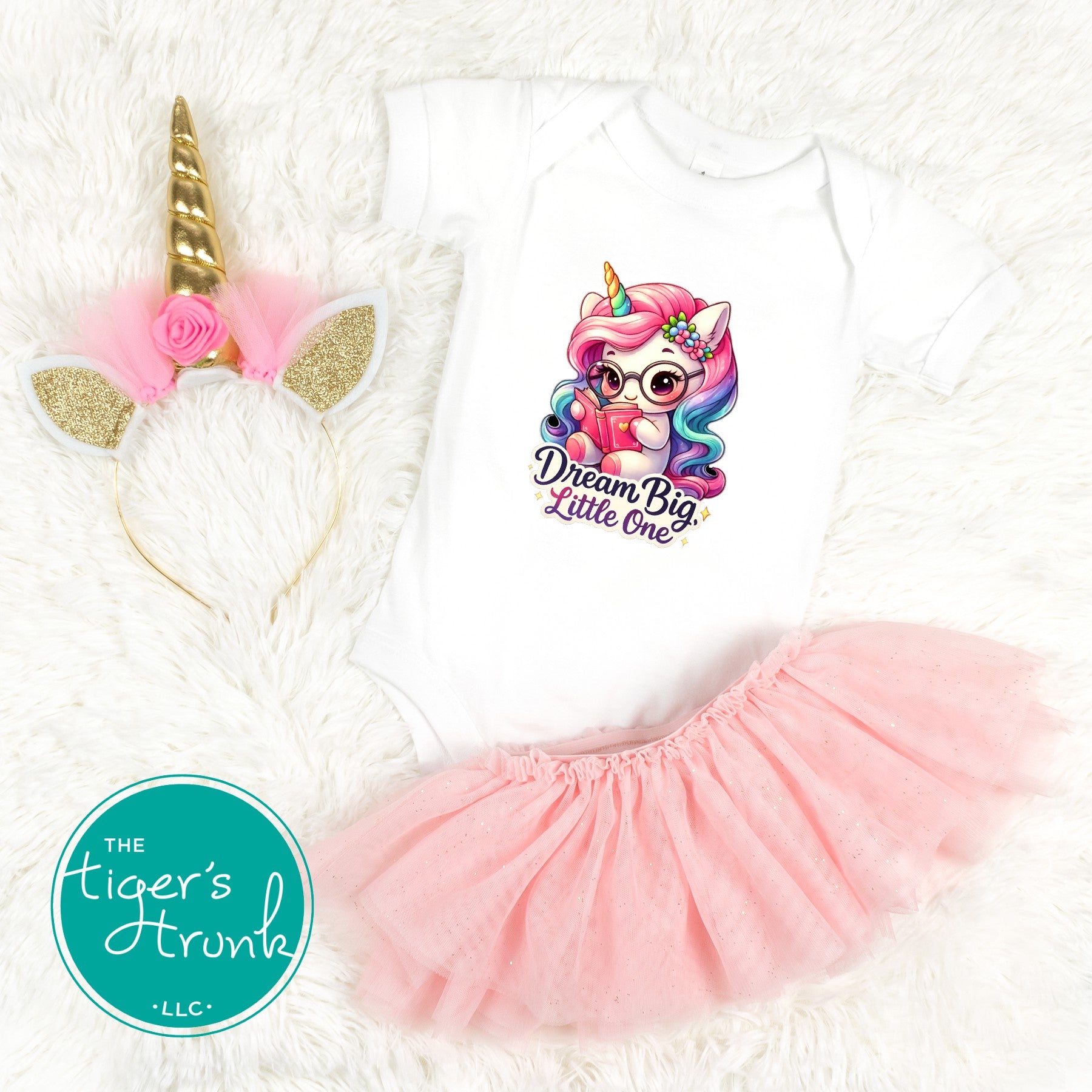 Infant white bodysuit on a white fur background with a colorful unicorn graphic that reads Dream Big Little One next to a tutu and a gold unicorn headband.