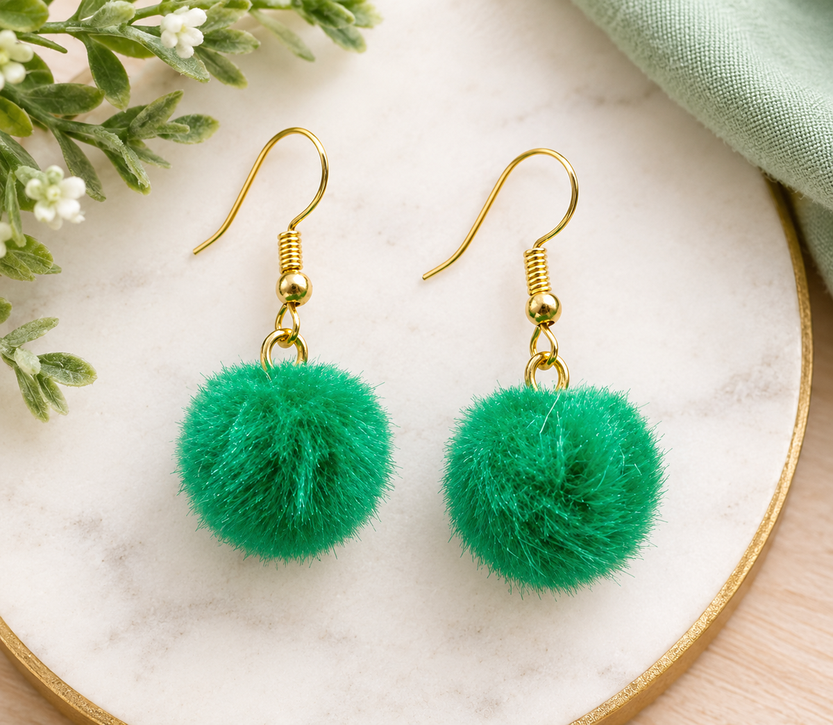 Emerald green fluffy pom pom dangle earrings with gold-tone fishhook ear wires displayed on a white marble tray, perfect for school spirit, game day, and St. Patrick’s Day.