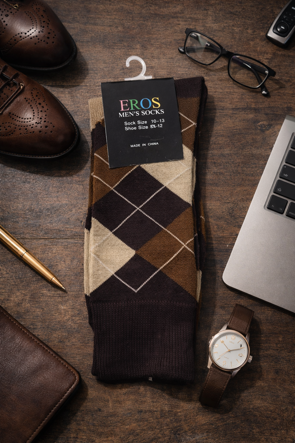 EROS men’s argyle crew socks in brown, tan, and cream diamond pattern with retail hang tag showing sock size 10–13 and men’s shoe size 8½–12, styled in a professional desk flatlay with leather shoes, glasses, pen, laptop, wallet, and wristwatch on a wood surface.