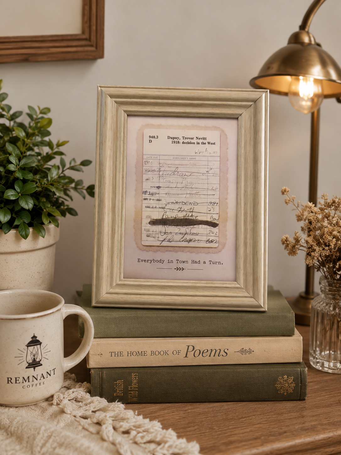 A vintage-style framed print featuring handwritten musical notation and the poem 'Everybody Is Too Idle to Be a Thief' displayed on a stack of books on a wooden table, styled with a mug, plant, lamp, and dried flowers.