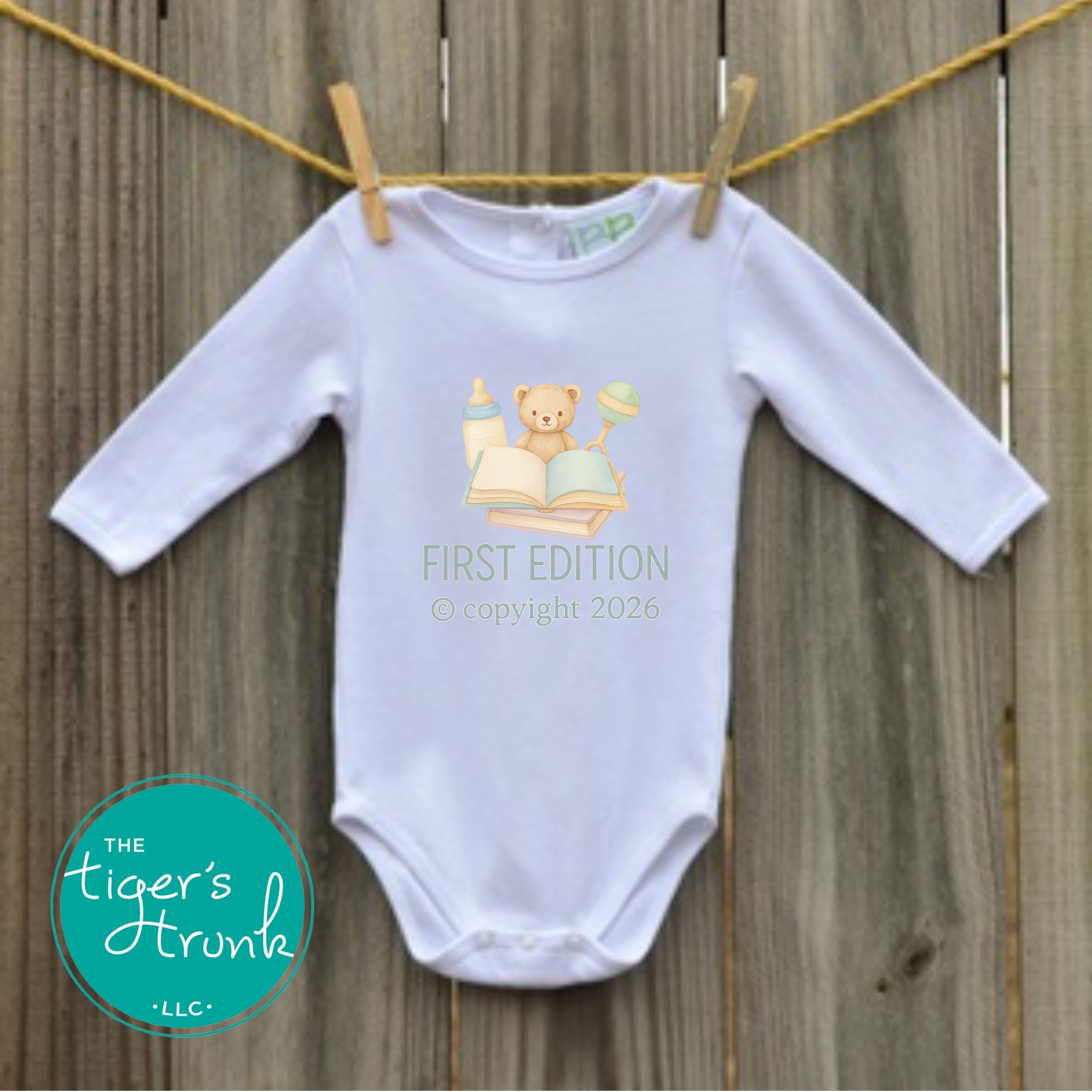 White long-sleeve baby bodysuit hanging on a clothesline with pastel illustration of a teddy bear, book, bottle, and rattle that reads First Edition.