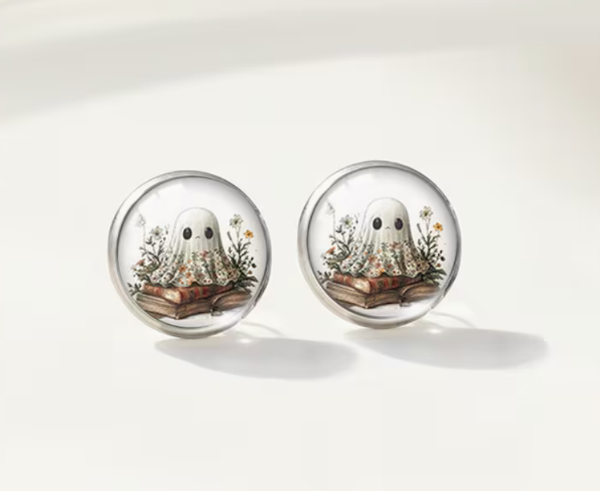 Round glass stud earrings featuring a cute white ghost sitting on vintage books surrounded by delicate wildflowers