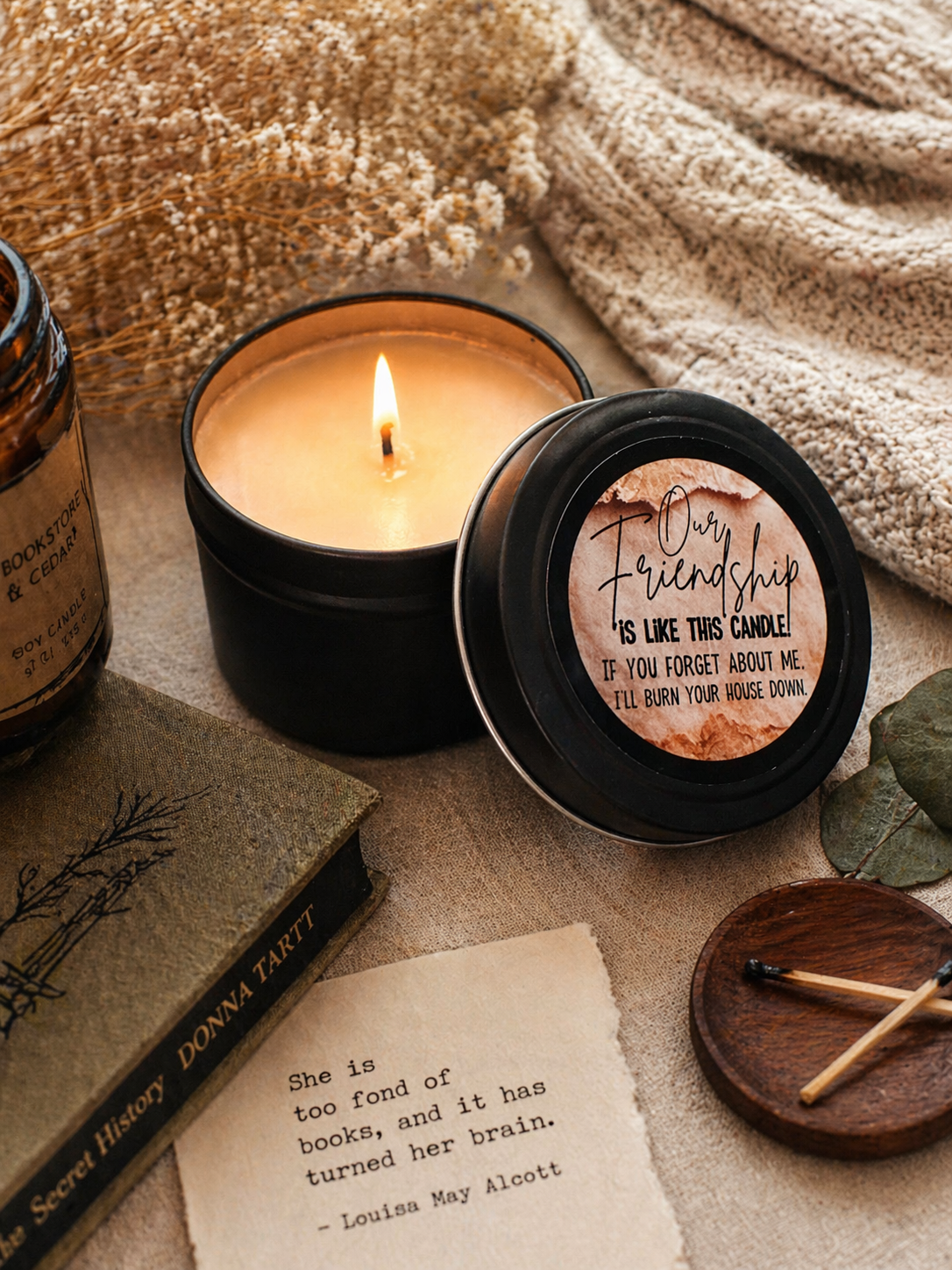 Candle in a black container with a humorous label, surrounded by books and a matchbox on a textured surface.