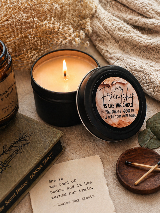 Candle in a black container with a humorous label, surrounded by books and a matchbox on a textured surface.