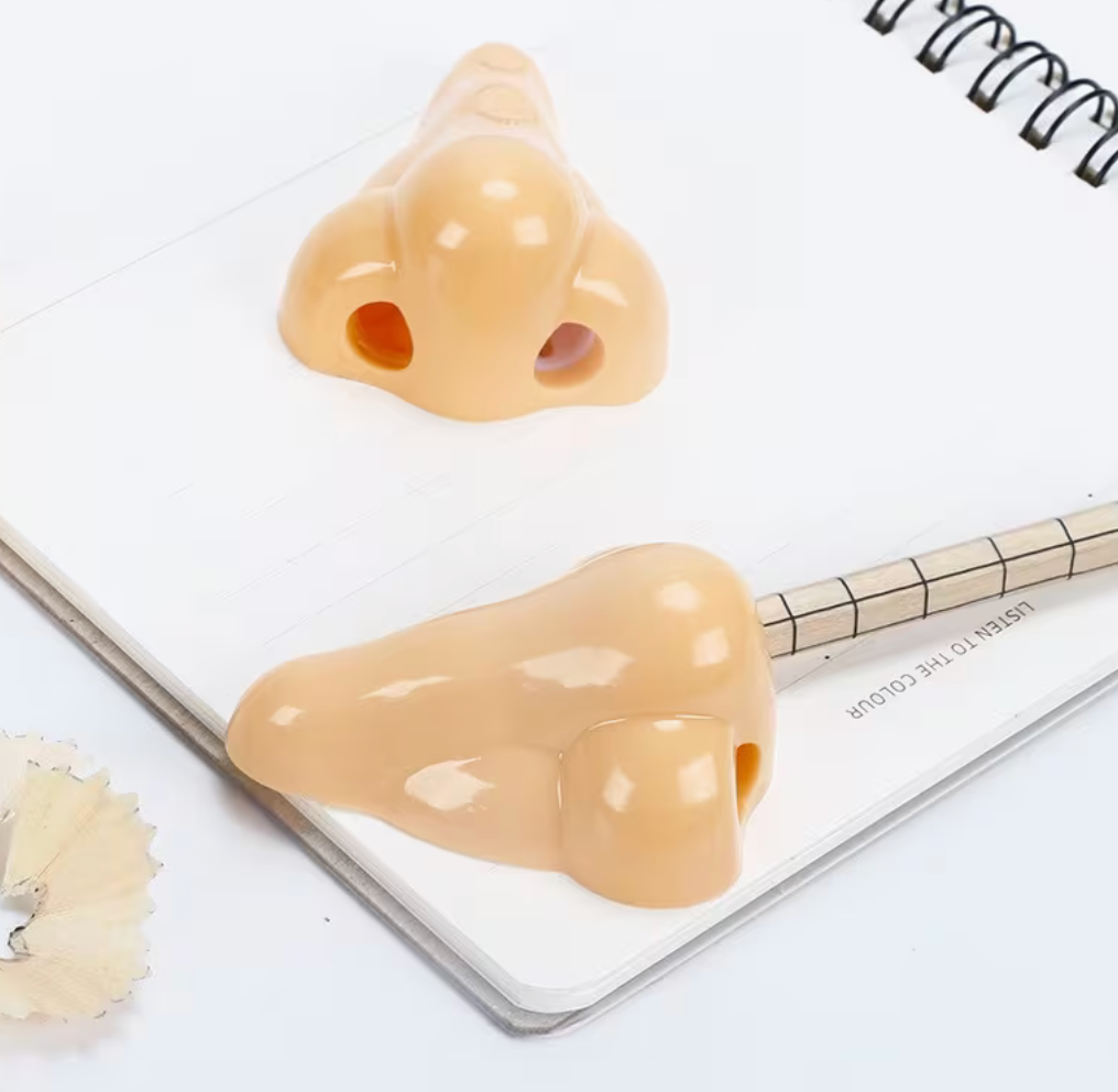 A novelty pencil shaped like a realistic peach-colored female vulva rests on an open spiral notebook, with a second matching pencil visible further back.