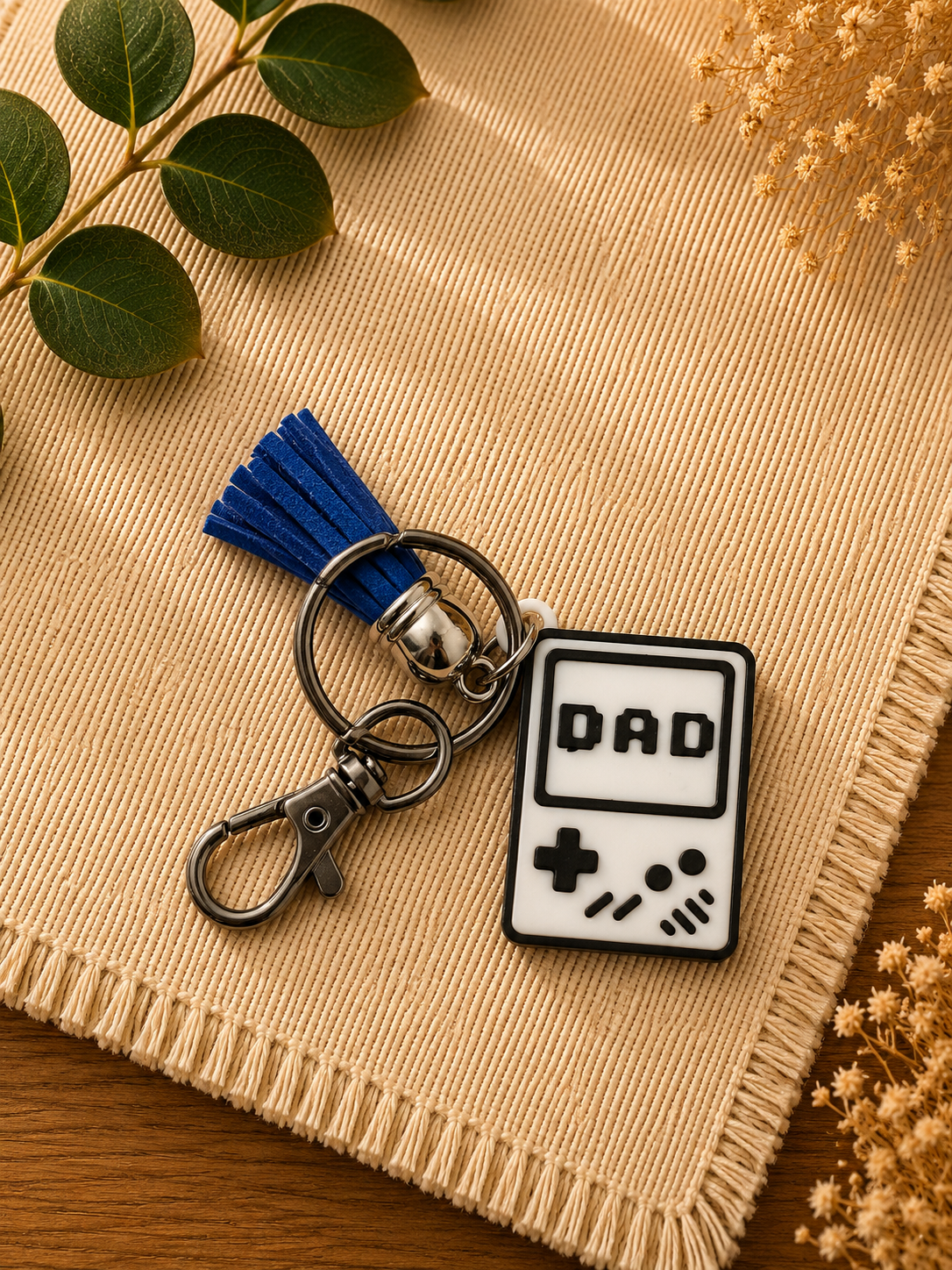 Retro handheld game console inspired dad keychain with blue tassel displayed on neutral woven fabric background