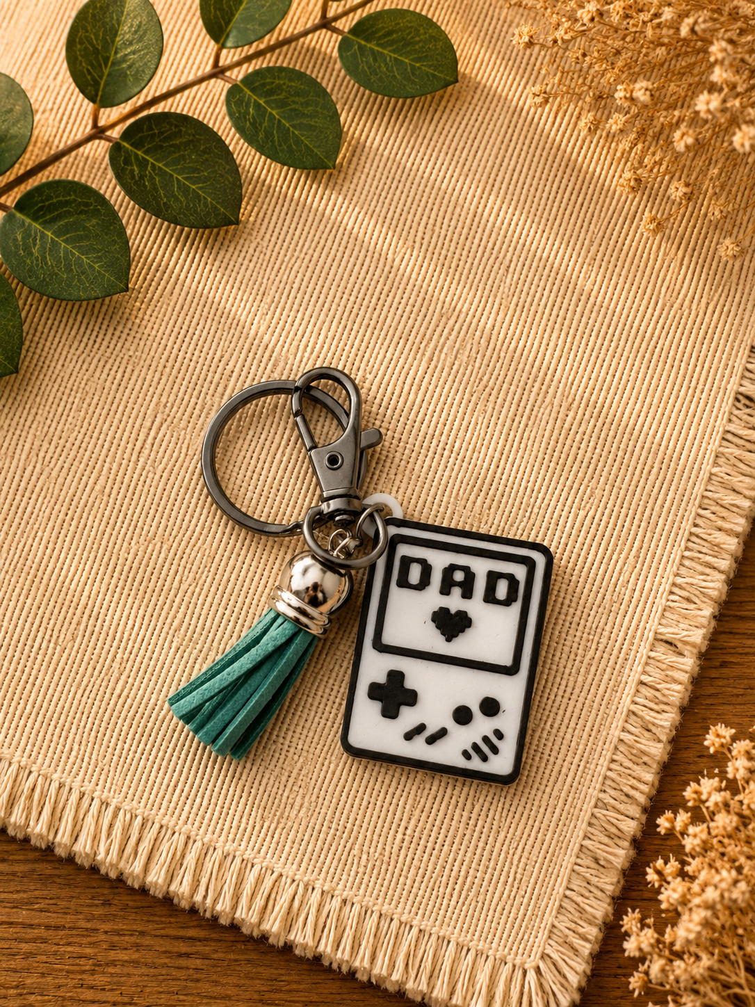 White and black gamer dad handheld console keychain with green tassel and lobster clasp on beige textured backdrop