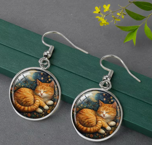 Cat-themed earrings on a gray surface with green leaves in the background
