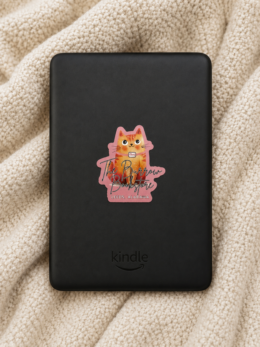 Pink Ginny the bookstore cat die cut sticker featuring The Burrow Bookstore logo displayed on a Kindle against a cozy cream blanket background.