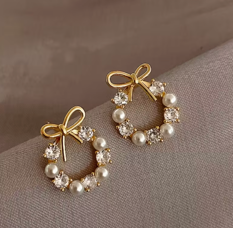 Golden-tone bow and pearl wreath earrings displayed on soft gray fabric, showing faux diamonds and delicate bow detail.