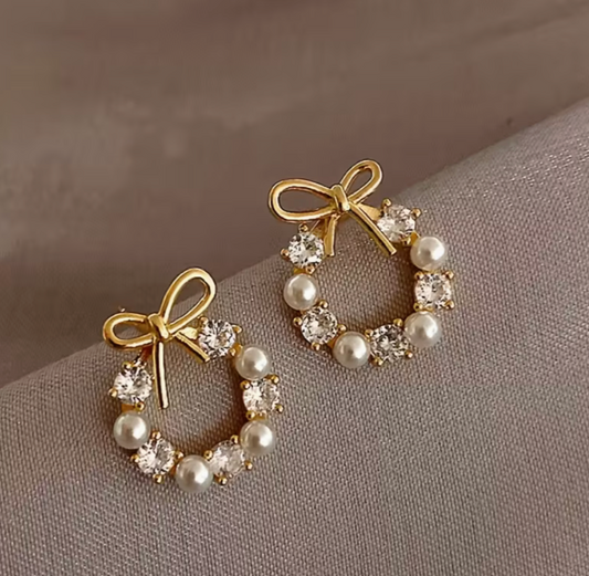 Golden-tone bow and pearl wreath earrings displayed on soft gray fabric, showing faux diamonds and delicate bow detail.