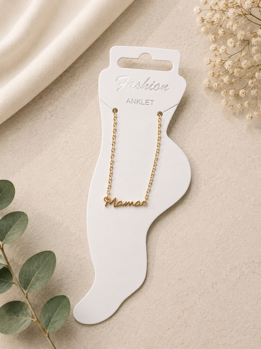 Gold mama anklet displayed on a foot-shaped jewelry card against an elegant neutral flat lay background with soft fabric, greenery, and delicate floral accents