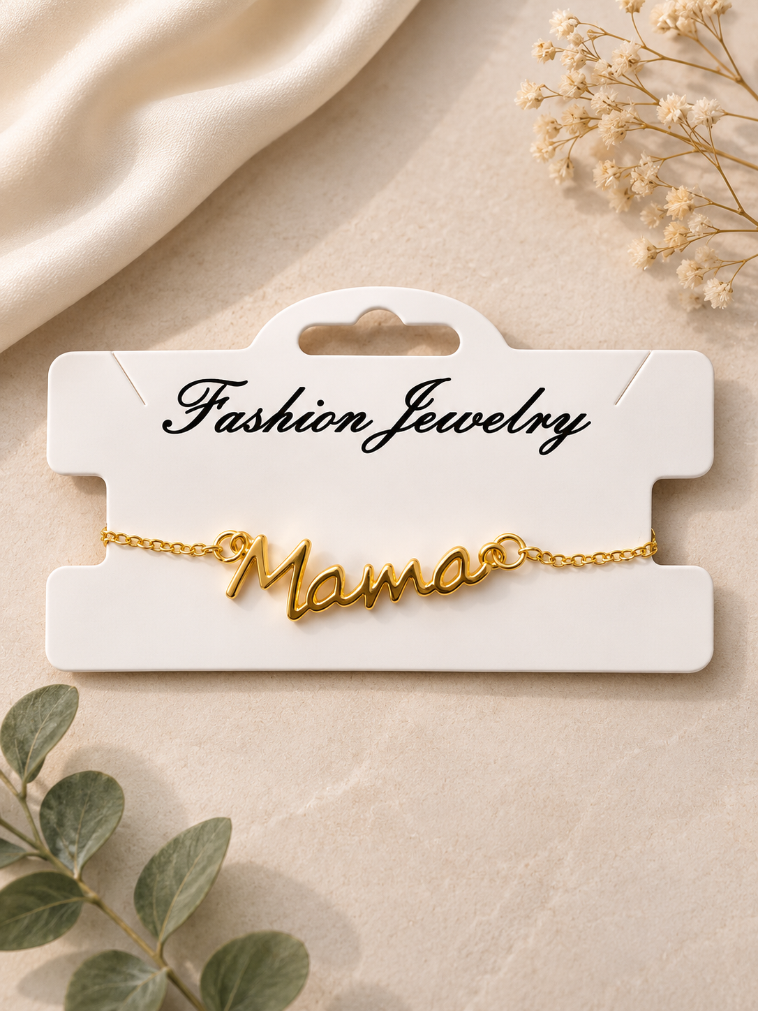 Gold mama script bracelet displayed on a white jewelry card against an elegant neutral flat lay background with soft fabric, greenery, and delicate floral accents