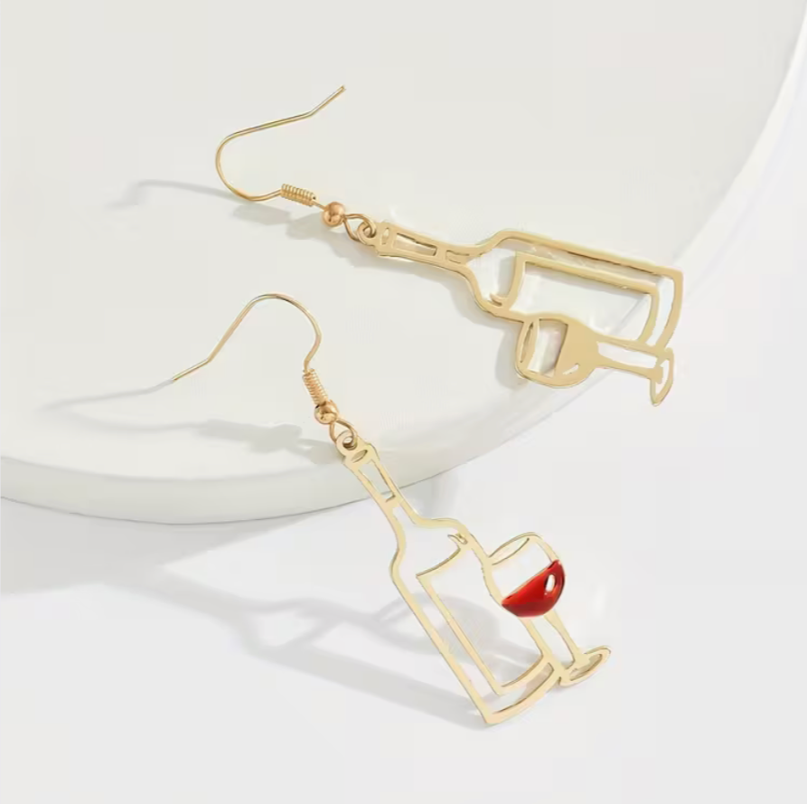 Flatlay of gold hollow-out wine bottle and wine glass earrings on a white surface.