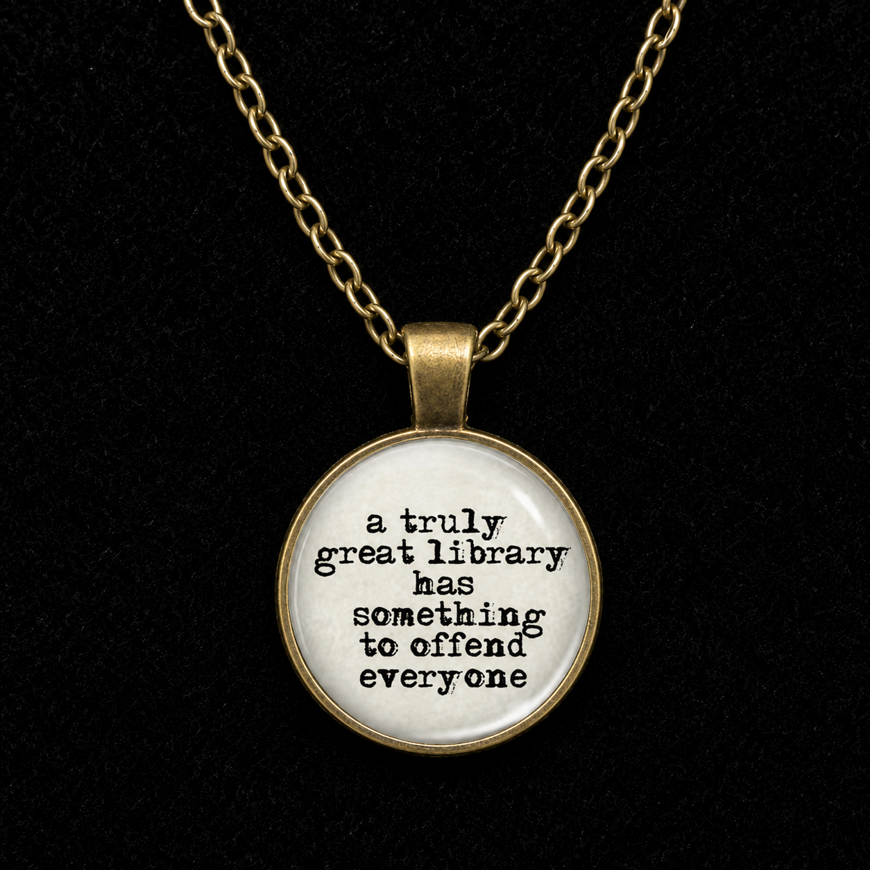 A round pendant necklace with a gold-tone chain and bezel, featuring a white face with black text that reads, 'a truly great library has something to offend everyone.' The pendant is shown against a solid black background.