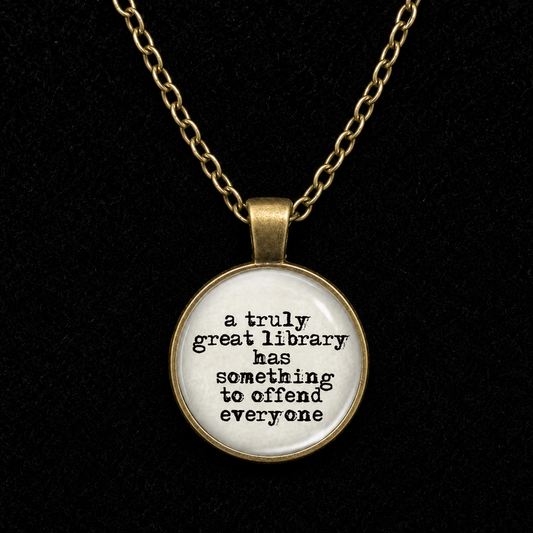 A round pendant necklace with a gold-tone chain and bezel, featuring a white face with black text that reads, 'a truly great library has something to offend everyone.' The pendant is shown against a solid black background.