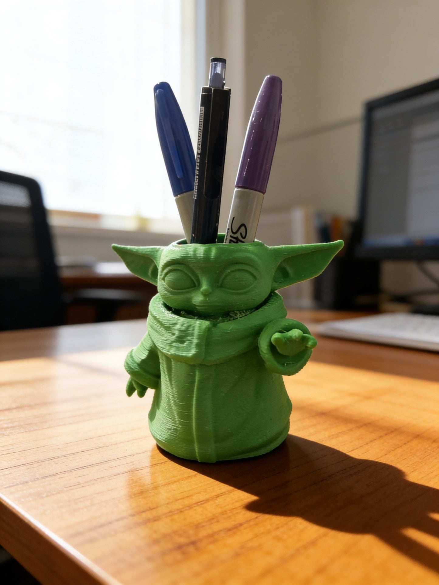 Green 3D printed character-inspired pen holder displayed on a wooden desk holding pens and markers, used as a desk organizer for office and school supplies.