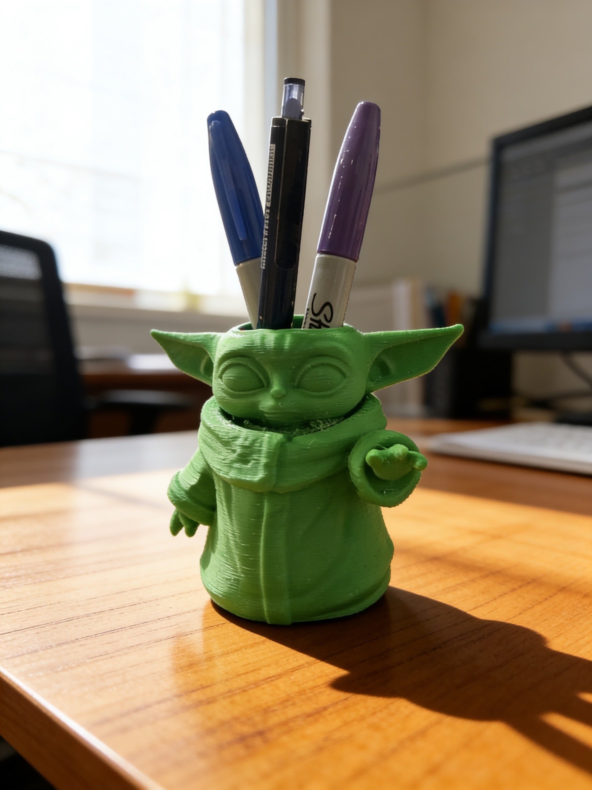 Green 3D printed character-inspired pen holder displayed on a wooden desk holding pens and markers, used as a desk organizer for office and school supplies.