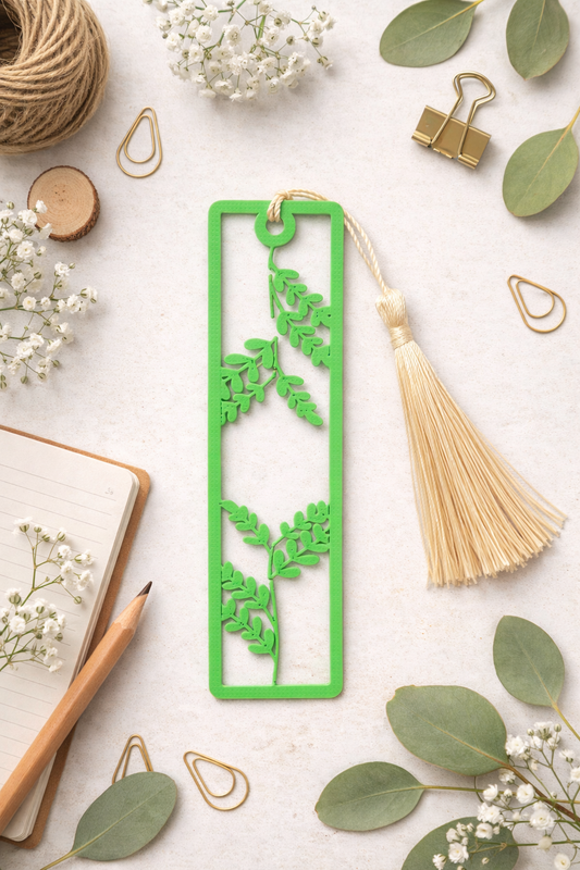 Green leaf-patterned bookmark with tassel on a white surface with flowers and stationery items.