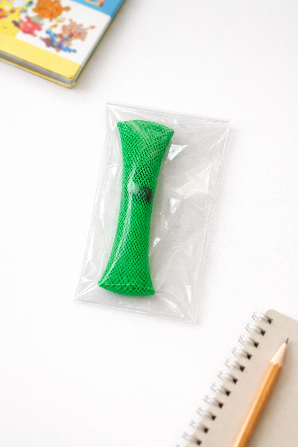 Green braided mesh marble fidget toy in clear plastic packaging on white desk with notebook, pencil, and colorful book in background.