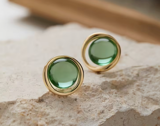 Green glass stone stud earrings with round gold-tone settings displayed on a neutral stone background