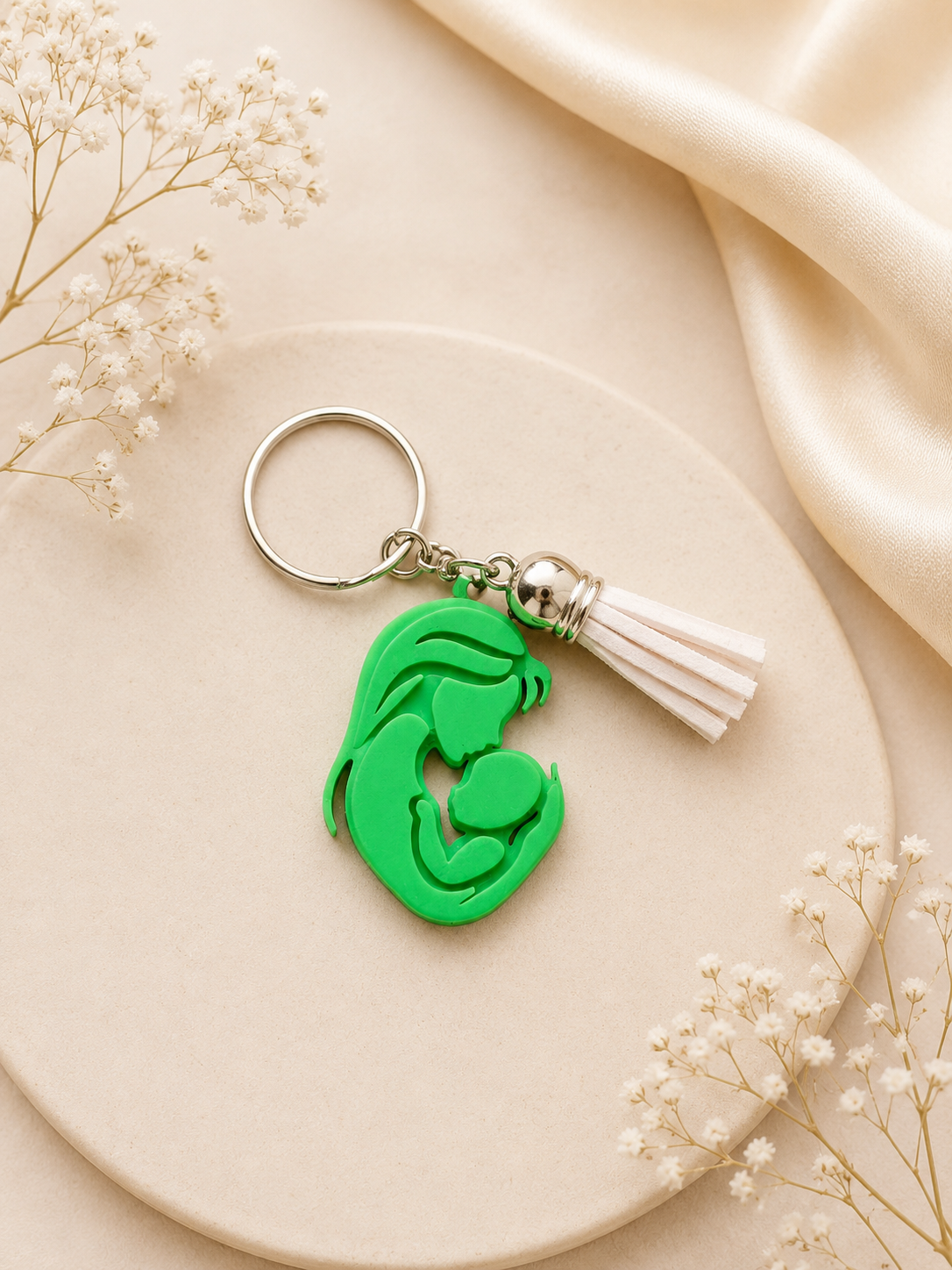 Green mother and baby silhouette keychain with white tassel displayed on a soft neutral flat lay background with delicate floral accents and satin fabric