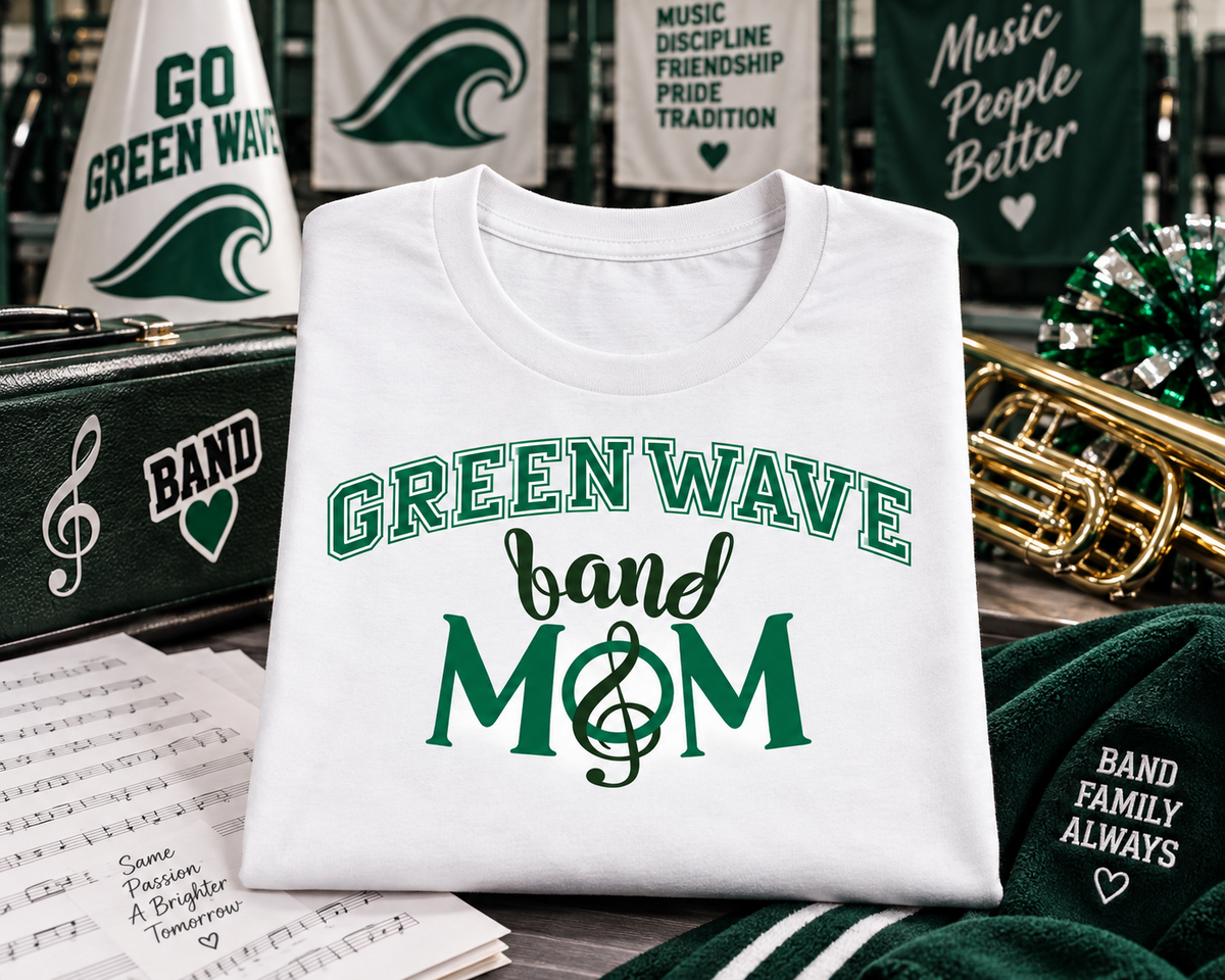 A folded white graphic T-shirt is displayed in a music-themed setting with green and white band apparel, sheet music, a brass instrument, and decorative props. The shirt reads “GREEN WAVE band MOM” in green lettering with a treble clef.