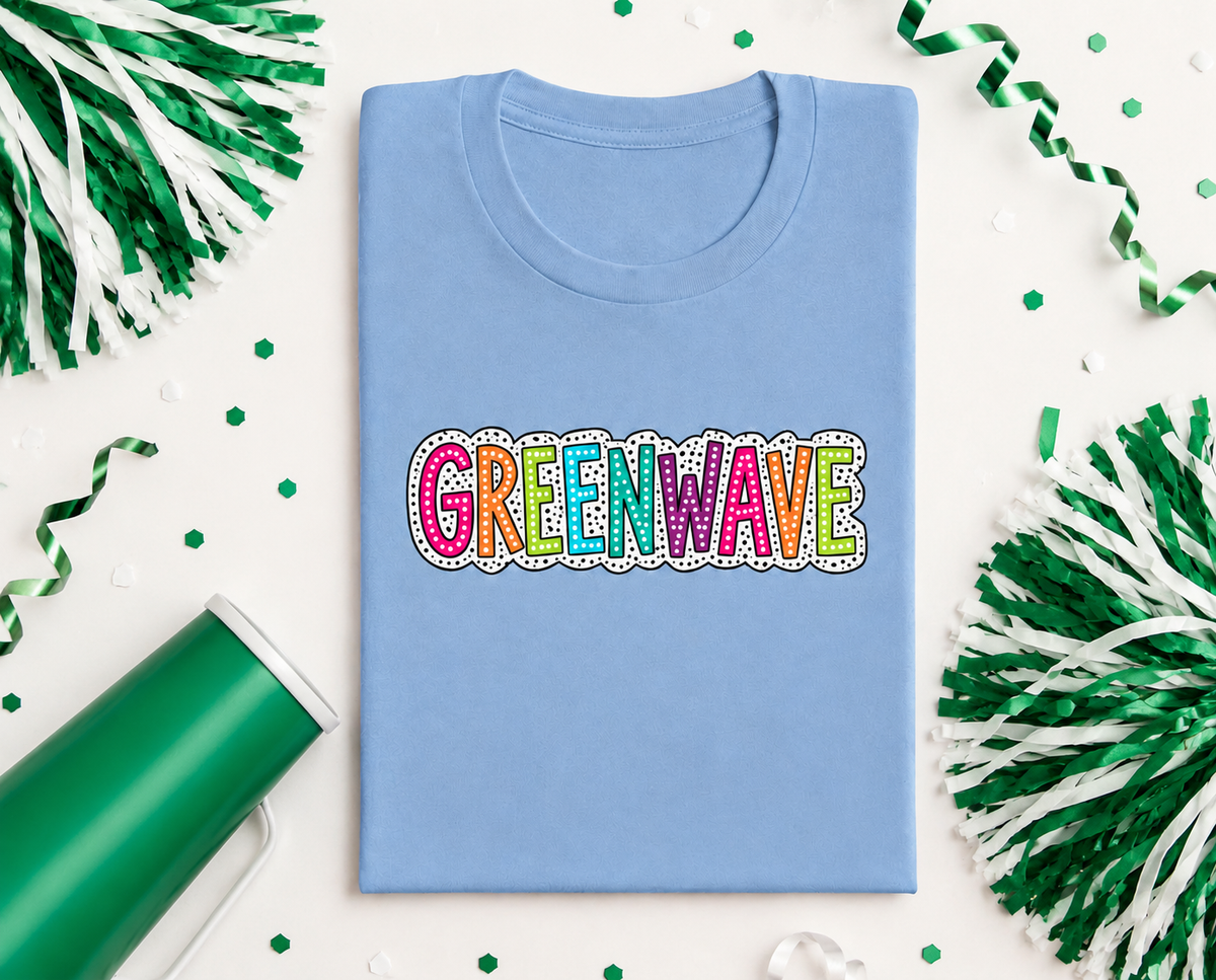 A folded light blue t-shirt with the word 'GREENWAVE' printed across the chest in colorful, bubbly letters outlined in white. The shirt is styled in a flat-lay with green and white pom-poms, green streamers, and green confetti on a white background.