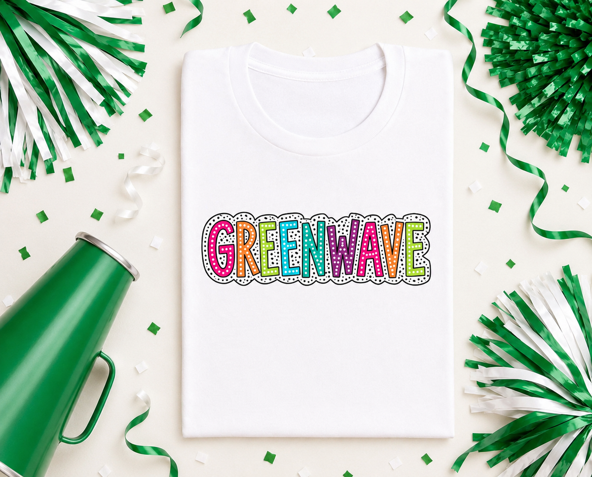 A white crewneck t-shirt laid flat on a light cream background, decorated with green and white pom-poms, streamers, and confetti. The shirt features a colorful graphic spelling 'GREENWAVE' in rainbow-colored letters with a black and white dotted border.