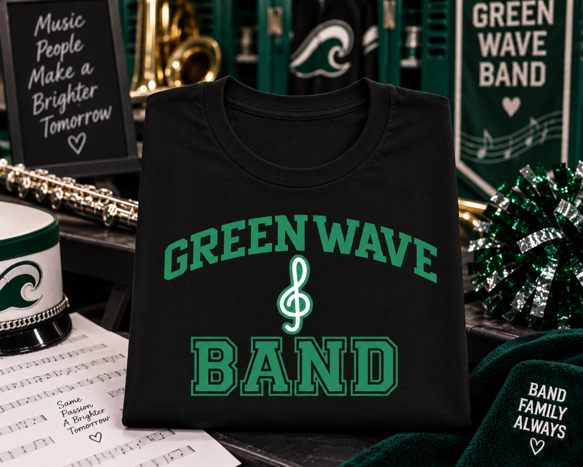 A black folded T-shirt with 'GREEN WAVE BAND' printed in green and white lettering, displayed on a table with music-themed props including a brass instrument, sheet music, a cheer pom-pom, and green signs.