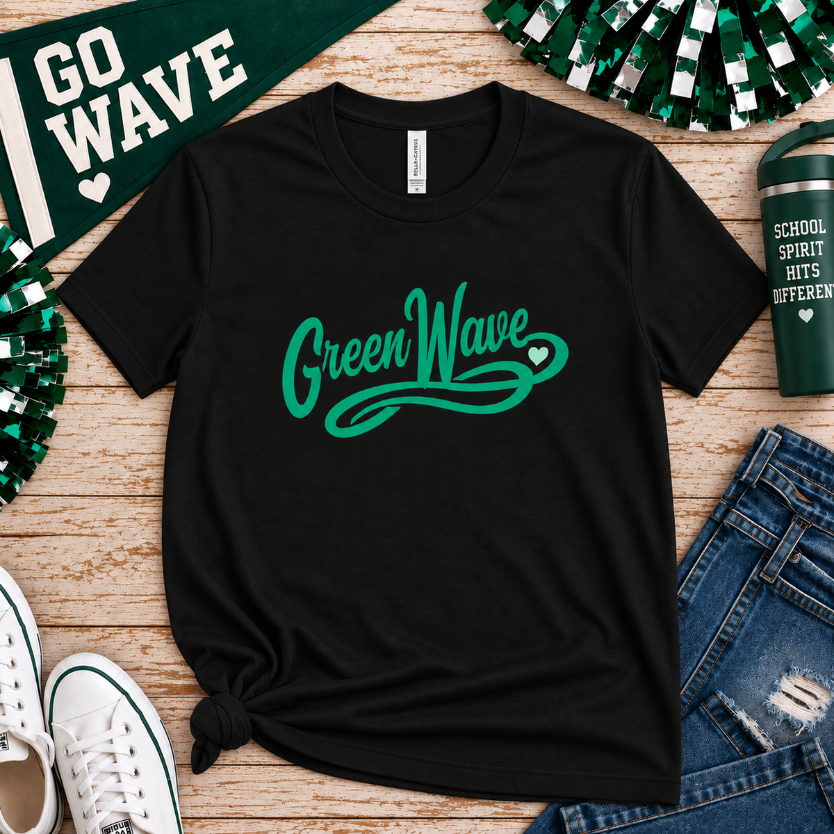 A black short-sleeve graphic T-shirt laid flat on a wood surface, featuring the words "Green Wave" in green script across the chest. The shirt is styled with a green pennant, pom-poms, white sneakers, blue jeans, and a green tumbler.