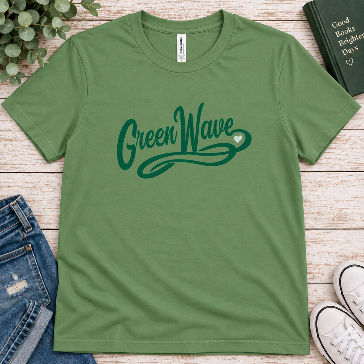 A green crewneck t-shirt with 'Green Wave' printed in dark green script across the chest, styled in a flat lay with jeans, a book, sneakers, and greenery on a light wood background.