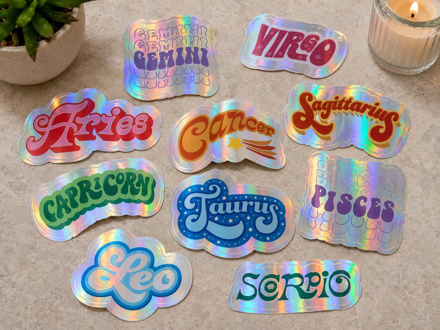 Flat lay of holographic zodiac sign die cut stickers scattered across a countertop, featuring Aries, Taurus, Gemini, Cancer, Leo, Virgo, Scorpio, Sagittarius, Capricorn, and Pisces designs with colorful retro lettering and iridescent shine.