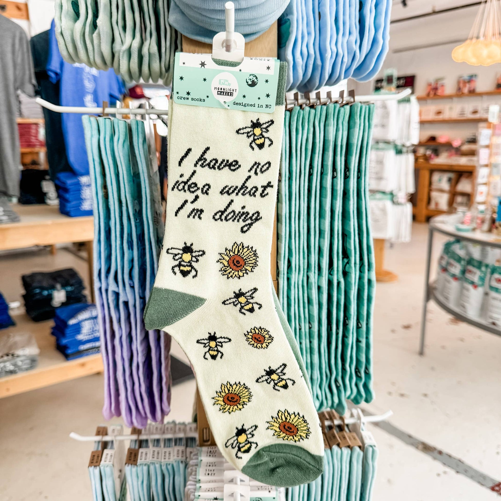 I have no idea what I’m doing novelty crew socks with bee and sunflower design hanging on a retail display rack, shown in store with Moonlight Makers packaging and surrounding pastel socks visible.