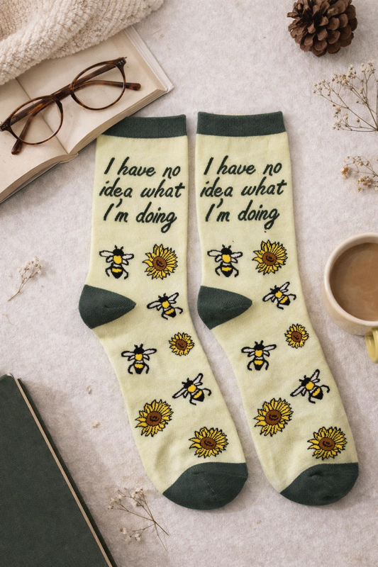 Flatlay of I have no idea what I’m doing novelty crew socks with bee and sunflower design in soft yellow and green, styled with an open book, reading glasses, coffee mug, pinecone, and cozy neutral background for a warm bookish gift look.