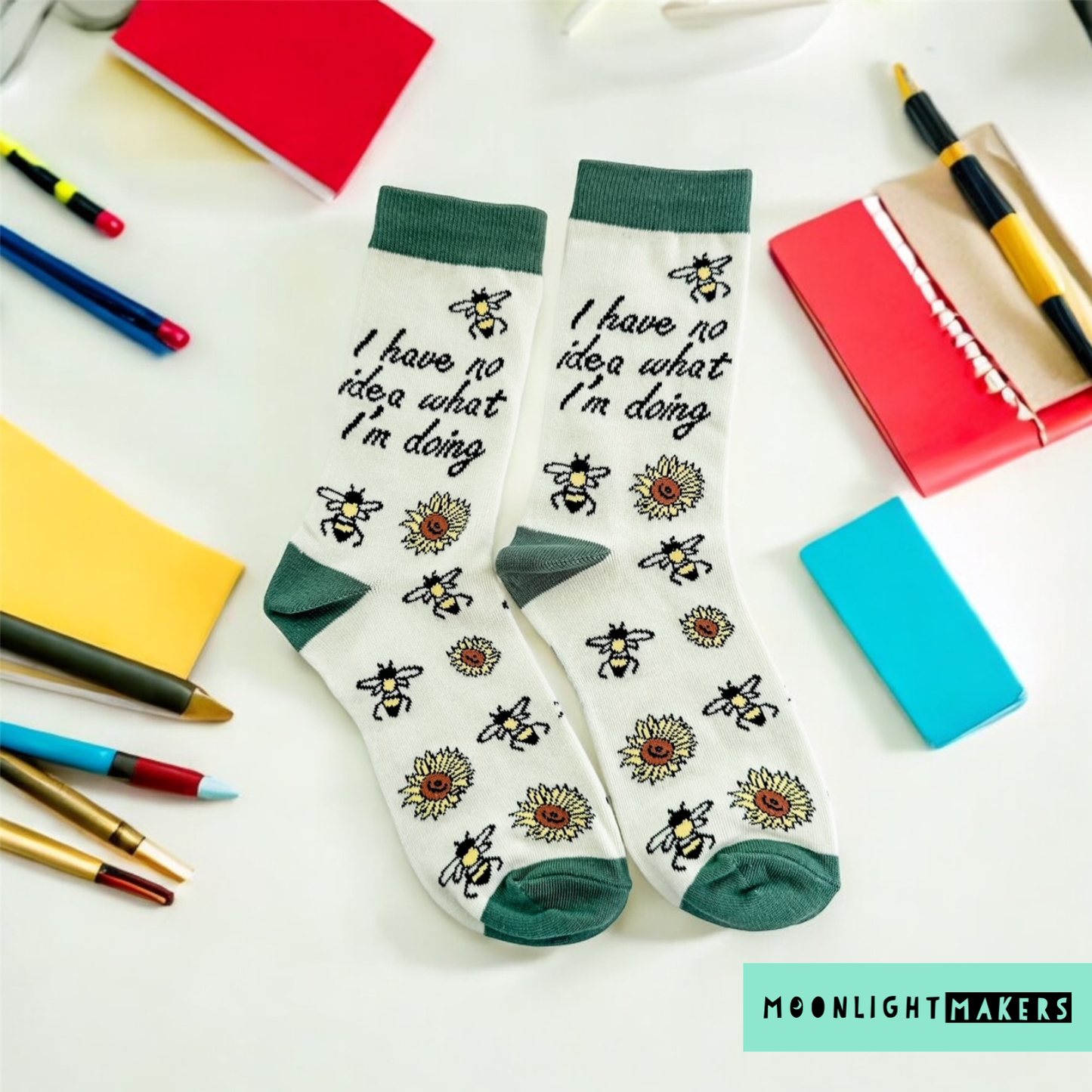 Flatlay of I have no idea what I’m doing novelty crew socks with bees and sunflowers on a cream background with green cuffs and toes, styled with colorful notebooks and pens, Moonlight Makers branding visible for product display.