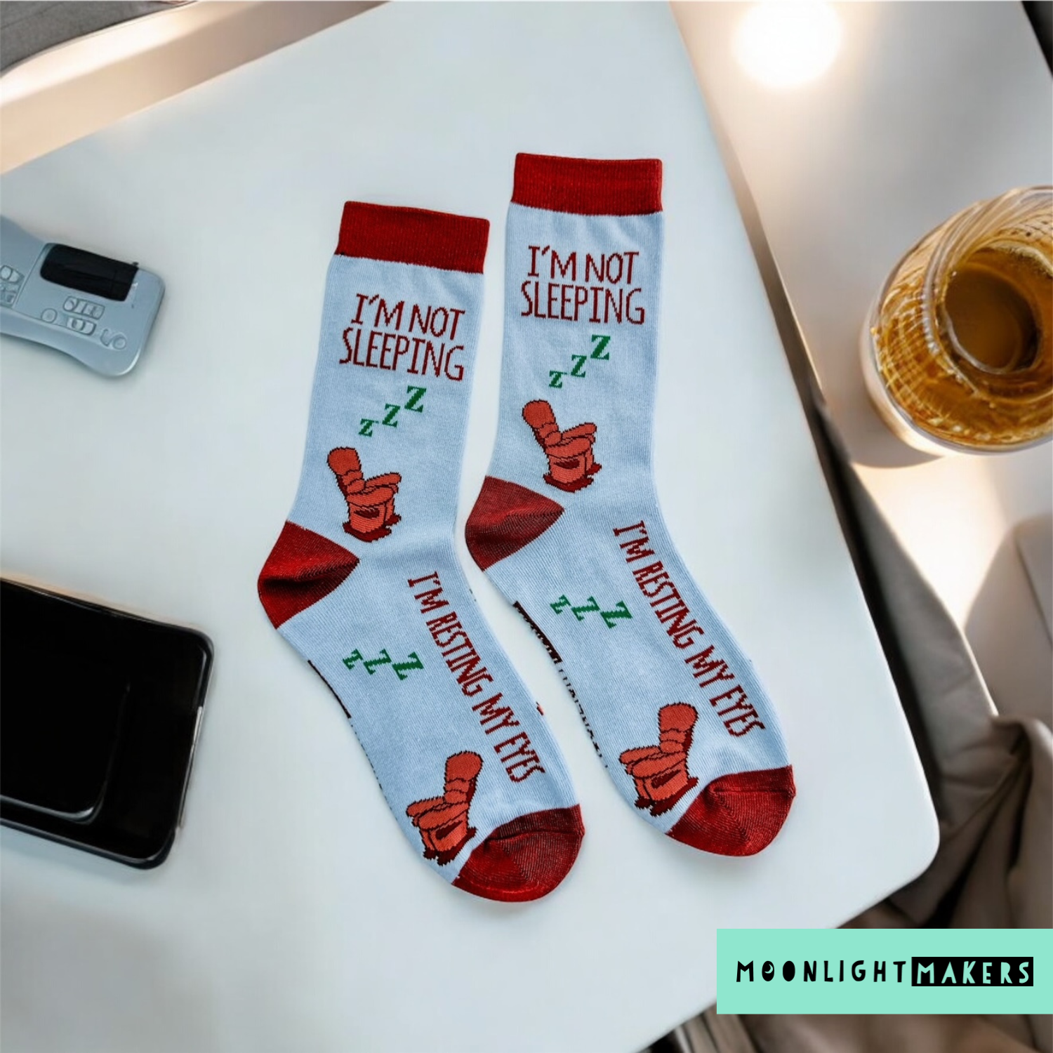 Flat lay of light blue novelty crew socks with recliner chair design and the phrases I’m not sleeping and I’m resting my eyes on a white desk with phone and drink nearby.