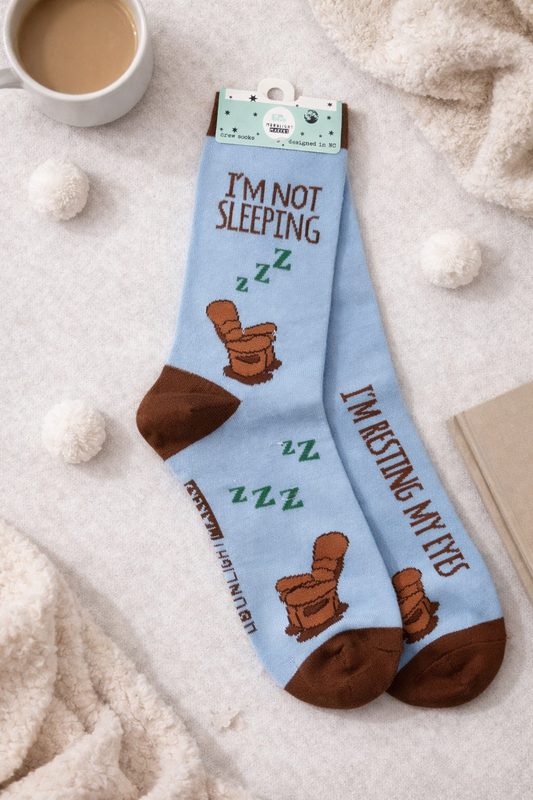 Light blue novelty crew socks that read I’m Not Sleeping and I’m Resting My Eyes, featuring a brown recliner chair and green ZZZ design on a cozy flat lay background.