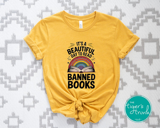 Yellow t-shirt with 'It's a Beautiful Day to Read Banned Books' text and rainbow design on a patterned background.