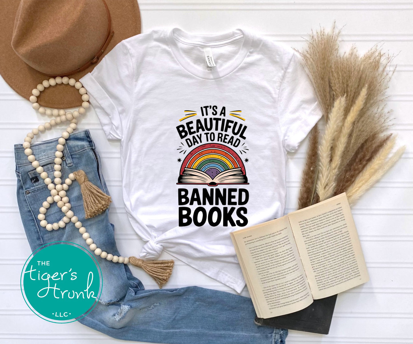 White t-shirt with 'It's a Beautiful Day to Read Banned Books' design, surrounded by denim jeans, a hat, and an open book.