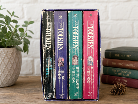 A set of four The Lord of the Rings book spines standing upright in a purple box, displayed on a wooden surface next to a potted plant and a stack of other books against a white brick wall.