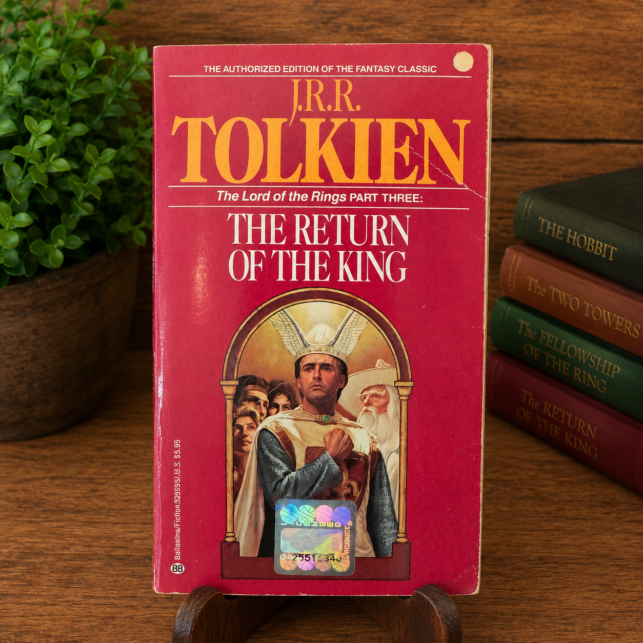 Vintage The Return of the King by J.R.R. Tolkien, Ballantine Books paperback, displayed upright on a wooden stand with a cozy bookshelf background. The front cover shows visible edge wear, a crease near the upper right corner, and the original holographic price sticker, accurately reflecting the book's used condition.