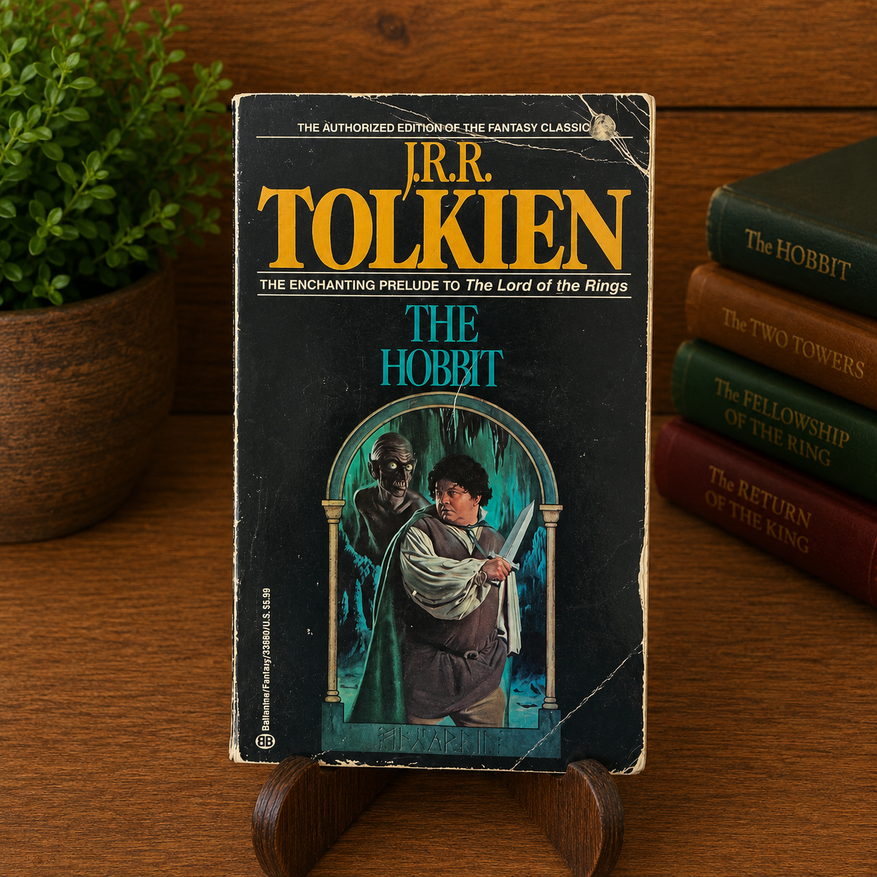 Vintage The Hobbit by J.R.R. Tolkien, Ballantine Books paperback, displayed upright on a wooden stand with a warm bookshelf background. The black front cover retains its authentic fair-condition wear, including edge wear, corner rubbing, surface creases, a small circular remainder hole punch near the upper-right corner, and visible creasing across the upper-right and lower-right portions of the cover.