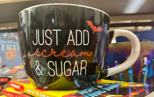 Black mug with 'Just Add Scream & Sugar' text on a stack of books.