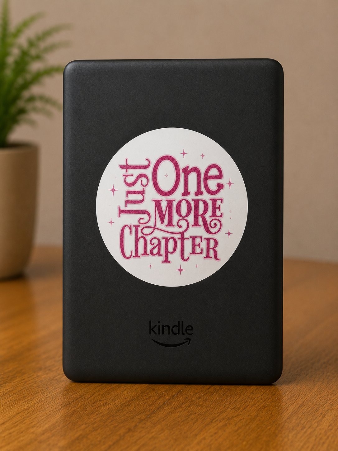 Pink Just One More Chapter reader sticker displayed on the back of a Kindle e-reader, featuring decorative typography and star accents on a white circular background.