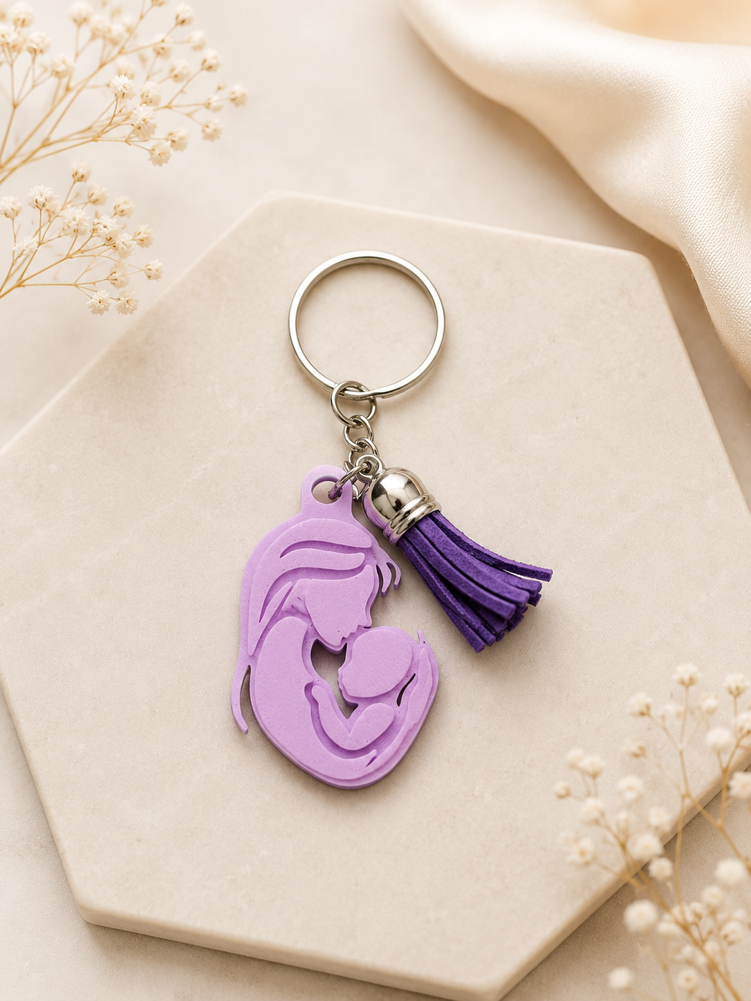 Lavender mother and baby silhouette keychain with purple tassel displayed on an elegant neutral flat lay background with soft floral accents and satin fabric