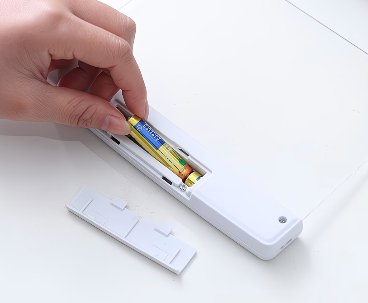 Hand inserting AAA batteries into the back compartment of a white LED flat panel reading light with the battery cover removed
